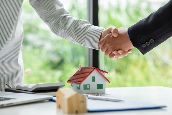 Two people shake hands, presumably over a real estate deal, with a table that contains a small model of a home beneath their hands.